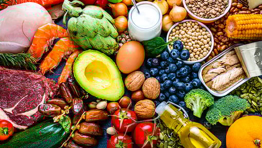 Assortment of fresh and healthy foods displayed on a surface, including avocado, blueberries, nuts, tomatoes, zucchini, broccoli, corn, olive oil, and more.