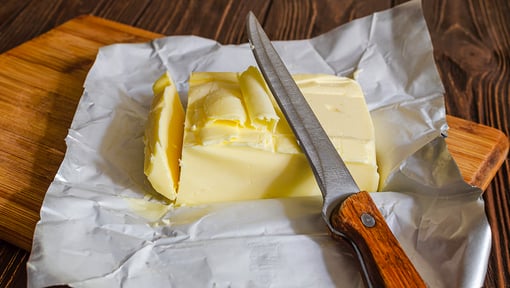 Block of butter partially sliced with a knife, resting on its wrapper atop a wooden cutting board.