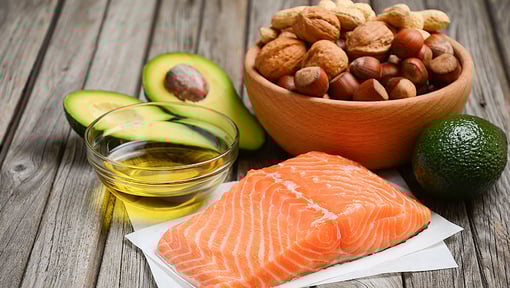 Close-up of healthy foods rich in good fats, including fresh salmon fillet, sliced avocado, a glass bowl of olive oil, and a wooden bowl filled with assorted nuts.