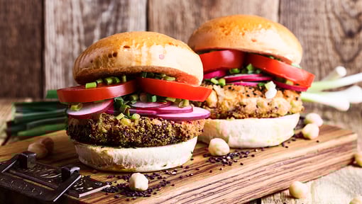 two gourmet veggie burgers on a wooden board, topped with fresh tomato slices, red onion rings, and chopped green herbs, with chickpeas scattered around