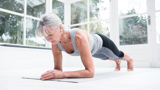 A woman holding an isometric forearm plank on a yoga mat in a bright indoor space with large windows.