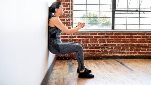 Fitness enthusiast performing a wall sit exercise in a gym setting, targeting legs and core strength.