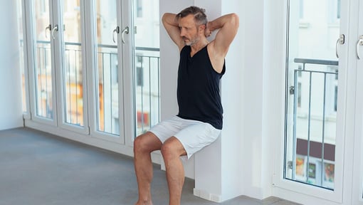 A man performing a joint-friendly exercise by doing a wall sit indoors, with arms bent and hands placed behind the head.