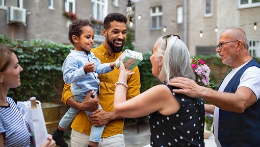 Group of people gathered outdoors in a courtyard, with one person holding a child and another offering a small gift, creating a warm and friendly interaction.