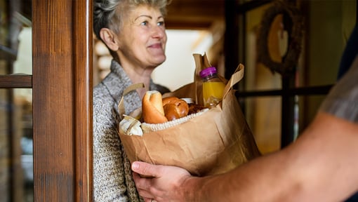 Person delivering a paper bag filled with groceries, including bread and juice, to someone at the door, showing an act of kindness.