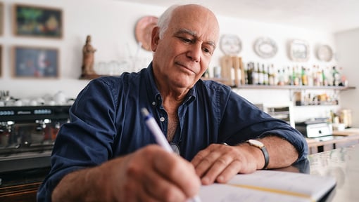 Person sitting at a table in a kitchen, writing in a notebook with a pen.