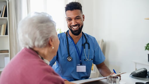 A doctor in blue scrubs smiling as he talks to an older patient across a desk in a clinical setting.