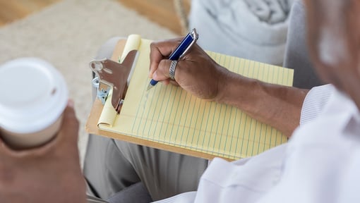 Person writing on a yellow notepad attached to a clipboard while holding a coffee cup.