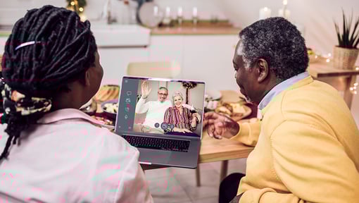 Two people sitting at a kitchen table looking at a laptop showing a video call with two individuals waving