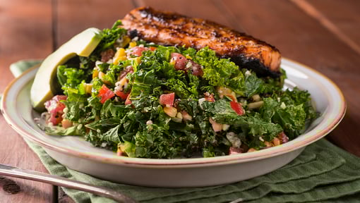 Plate of fresh kale salad mixed with diced vegetables, served alongside a grilled salmon fillet and avocado slices