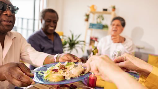 A group of people sharing a meal at a dining table, passing a plate of food in a warm, home setting that emphasizes communal eating and nourishment.