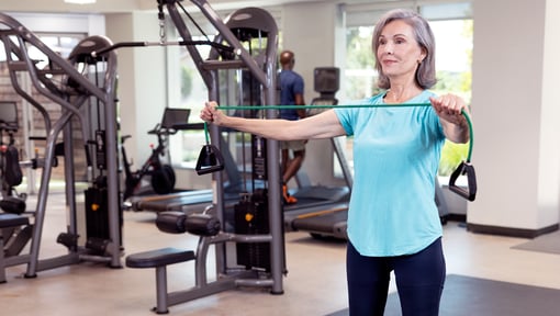 An older adult exercising in a gym, using resistance bands in front of strength-training equipment as part of a fitness routine.