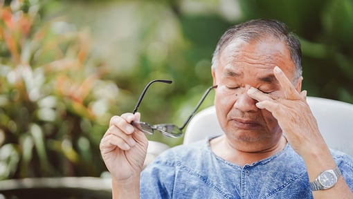 rson holding eyeglasses and touching the eye area while seated outdoors, illustrating eye discomfort or visual strain.