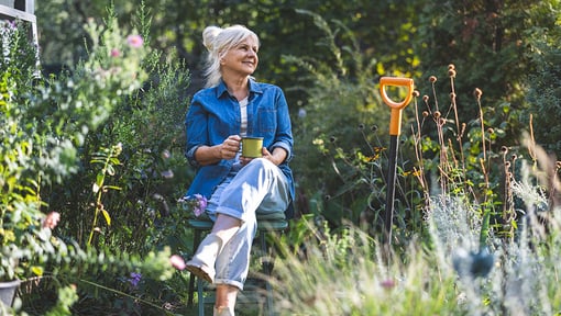 An older adult woman enjoys a slow living moment, sitting in a garden with a warm drink surrounded by greenery.
