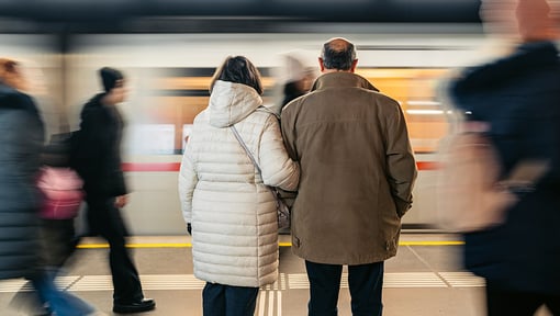Two older adults standing together on a subway platform as a train speeds past in motion blur.