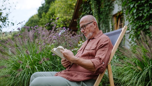 A person relaxes in a wooden chair, reading a book in a quiet garden with tall grasses and greenery.