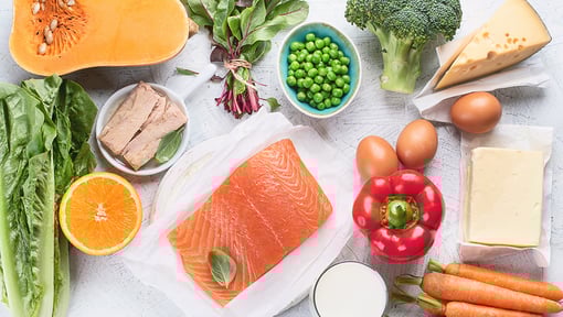 Assortment of whole foods rich in vitamins and minerals, including salmon, leafy greens, broccoli, peas, eggs, carrots, cheese, and citrus