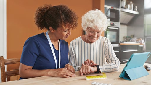 A caregiver and an older adult sitting at a kitchen table looking at a smart watch 