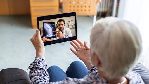 An older adult sits holding a tablet during a video call with a healthcare professional, who is pointing to a medical scan displayed on the screen.