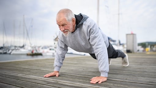 A man performing a plank-style exercise on a wooden dock near a marina.