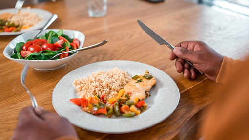 Person eating a balanced meal with salmon, rice, and sautéed vegetables at a wooden table.