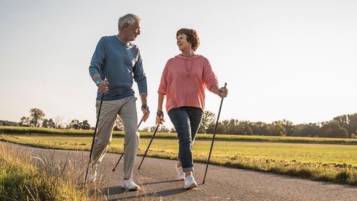  two older adults walking on a paved trail with walking poles. 