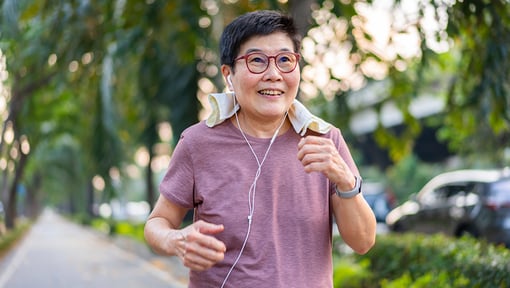 Older adult person walking along a sidewalk in a green neighborhood setting, wearing earbuds and athletic clothing.