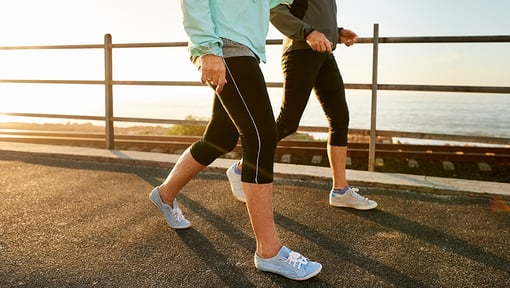Two individuals walking outdoors on a paved path near a waterfront, shown from the waist down, wearing athletic shoes and leggings.