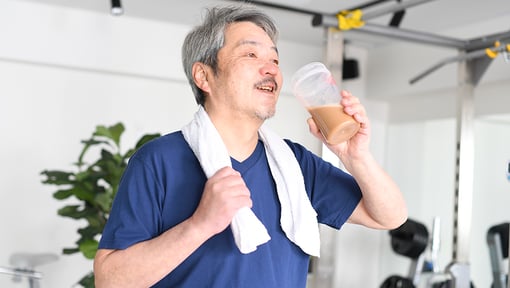Older adult in a gym setting drinking a creatine protein shake, with a towel draped over one shoulder 