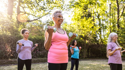 Group of older adults exercising outdoors in a park, each holding light dumbbells while performing standing strength‑training movements among trees