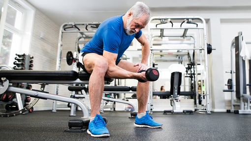 Older adult exercising in a gym, seated on a workout bench while performing a single‑arm dumbbell curl with weights nearby.