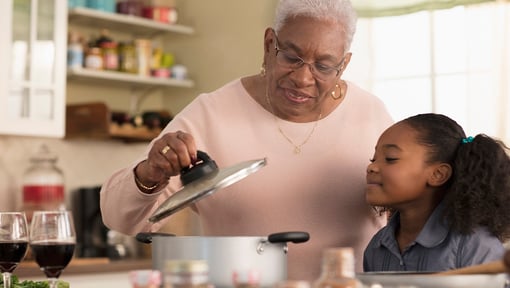 An older adult and a child stand at a kitchen counter, looking into a large pot as the older adult lifts the lid.