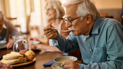 An older adult man sits at a dining table, eating soup from a bowl with a spoon. The table holds a plate of fruit, including bananas and apples, along with drinking glasses