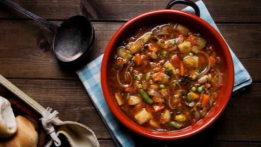 Overhead view of a rustic bowl of hearty vegetable soup filled with potatoes, carrots, green beans, peas, onions, and herbs in a rich broth
