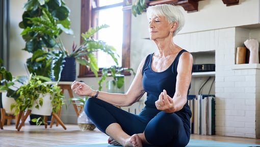 A older woman sits cross-legged on a yoga mat indoors, practicing seated meditation with eyes closed and hands resting on the knees.