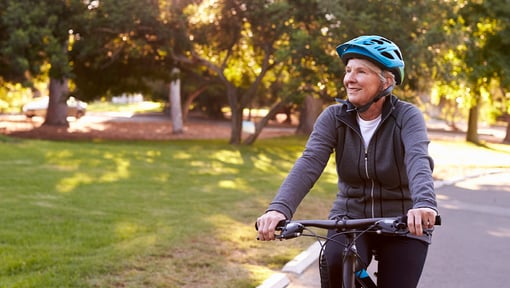 A woman rides a bicycle along a paved path in a park, wearing a helmet and a zip-up jacket.