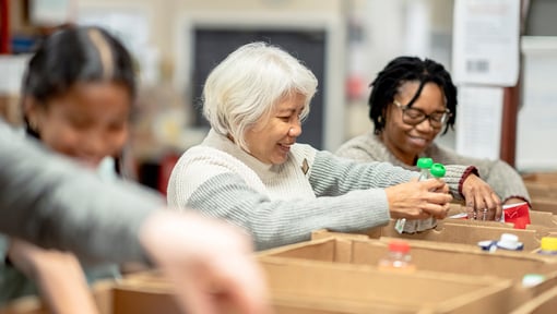 Three people are volunteering and working side by side at a table, placing small items into open cardboard boxes.