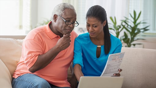 An older adult man and woman sit on a couch while looking at some paperwork and a computer. 