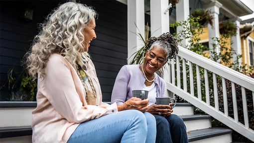 Two older adult women sit on the front steps of a house, enjoying some coffee together while laughing. 