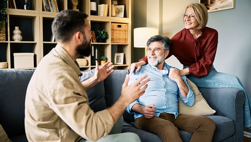 Three adults are gathered in a living room, with two people seated on a couch while another sits nearby, gesturing with hands as if explaining or discussing something. 