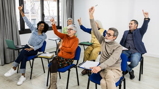 Several older adults seated in  classroom-style chairs, some are raising one hand, some have notebooks on their laps 