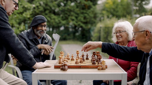 Several older adults seated around a small outdoor table, playing a game of chess.