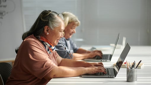 Two older adult women sit at a table with laptops in front of them during a continuing education course. 