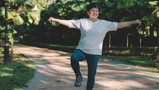 Older adult person balancing on one leg with arms extended on a walking path in a park, surrounded by trees and greenery