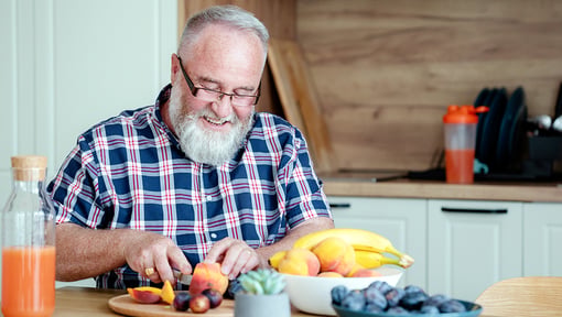 A man preparing fresh fruit in a kitchen, cutting peaches at a table with bananas, grapes, and other healthy foods nearby.