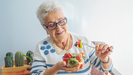 An older adult woman holding a bowl of fruit salad in one hand and in the other a fork stacked with fruit. 