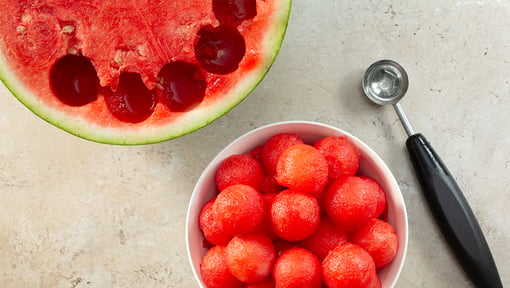 Overhead view of fresh watermelon, including a halved watermelon, a bowl of watermelon balls, and a melon baller tool on a countertop