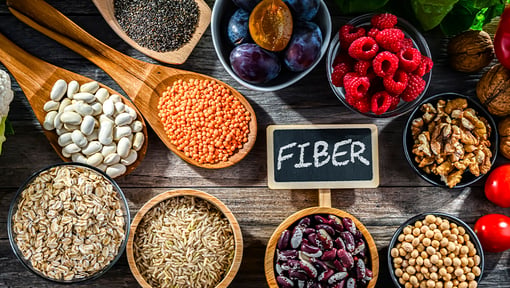 Assortment of high-fiber foods including oats, beans, lentils, brown rice, chia seeds, berries, walnuts, and soybeans arranged on a wooden table.