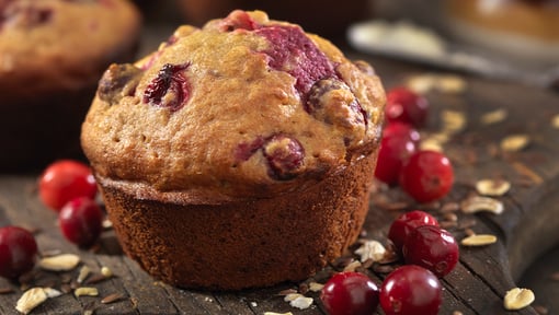 A freshly baked pumpkin cranberry muffin on a wood surface with whole cranberries and oats scattered around it. 