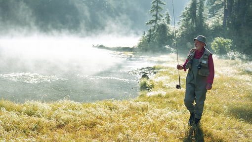 A man wearing outdoor gear and holding a fishing rod while standing beside a mist-covered lake bordered by trees and grassy shoreline.
